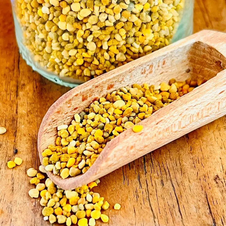 Scoopa de madeira com grânulos de pólen apícola amarelos e verdes ao lado de frasco em superfície de madeira