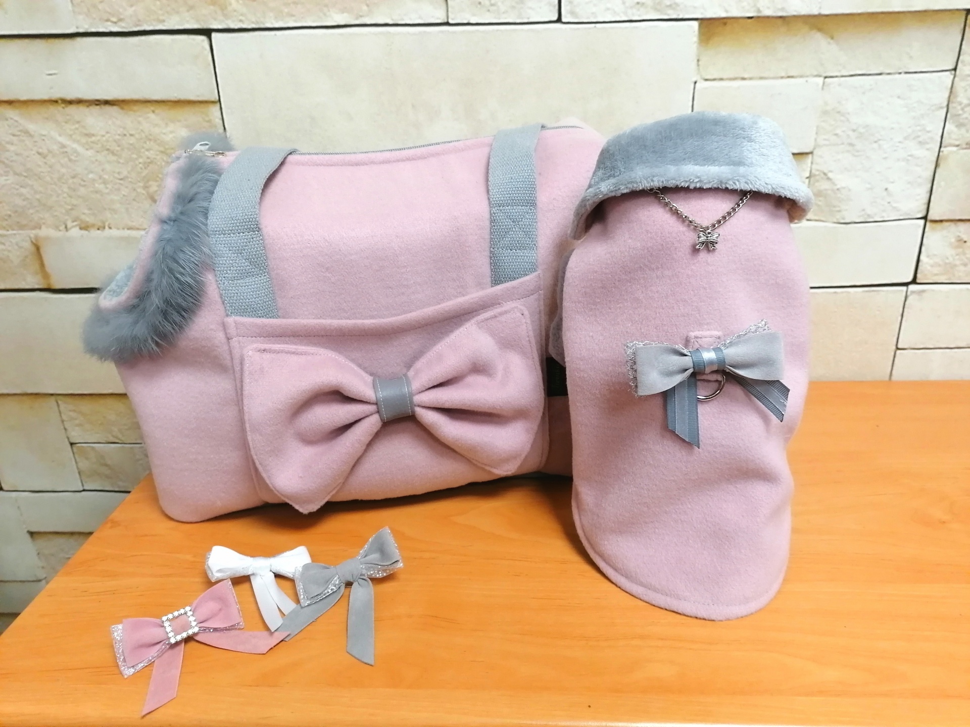 Bolsa e casaco para cão rosa com laços cinzentos e acessórios coloridos sobre mesa de madeira.