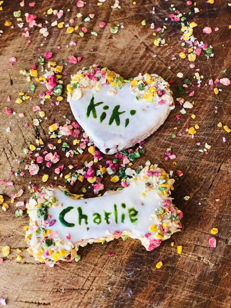 Biscoitos para cães em formato de osso e coração com nomes escritos e cristais coloridos sobre madeira.
