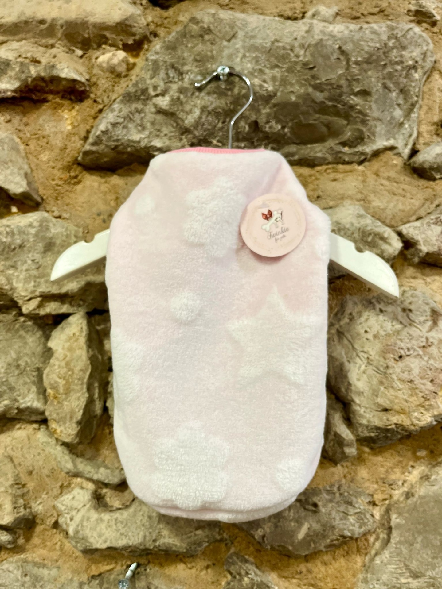 Roupa rosa clara para animal com padrão de estrelas e orelhas, pendurada em cabide contra parede de pedra.