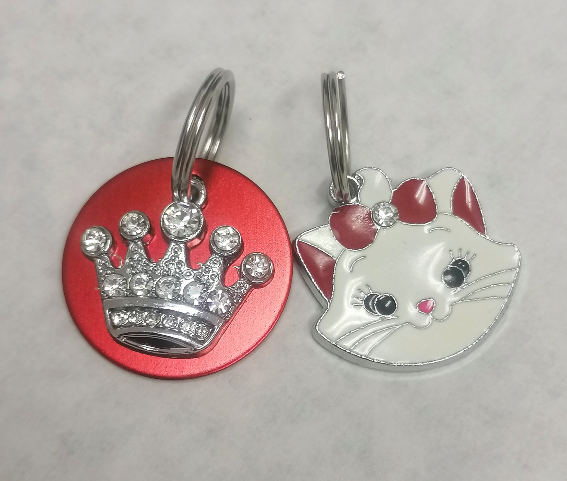 Dois porte-chaves metálicos, um redondo vermelho com coroa de cristais e outro em forma de gato branco com laços vermelhos.