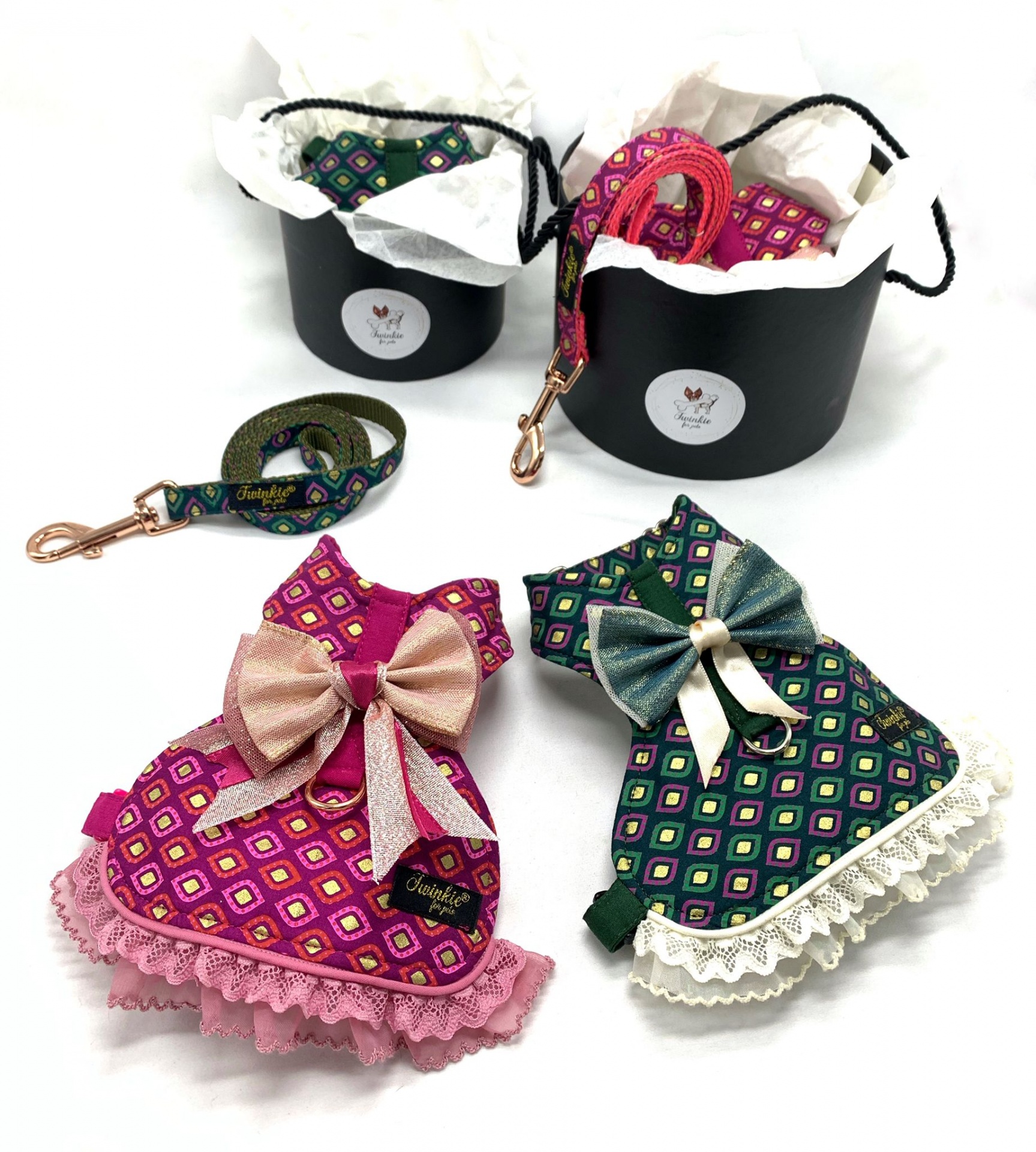 Dois vestidos e coleiras coloridos para animais, com padrão geométrico e rendas, exibidos junto a caixas pretas com tecido branco