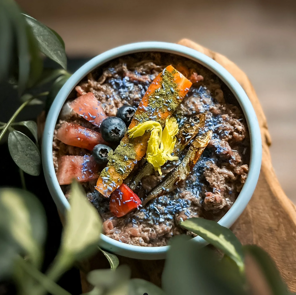 Prato azul com comida que inclui vegetais, mirtilos, sardinhas e flocos azuis em tábua de madeira