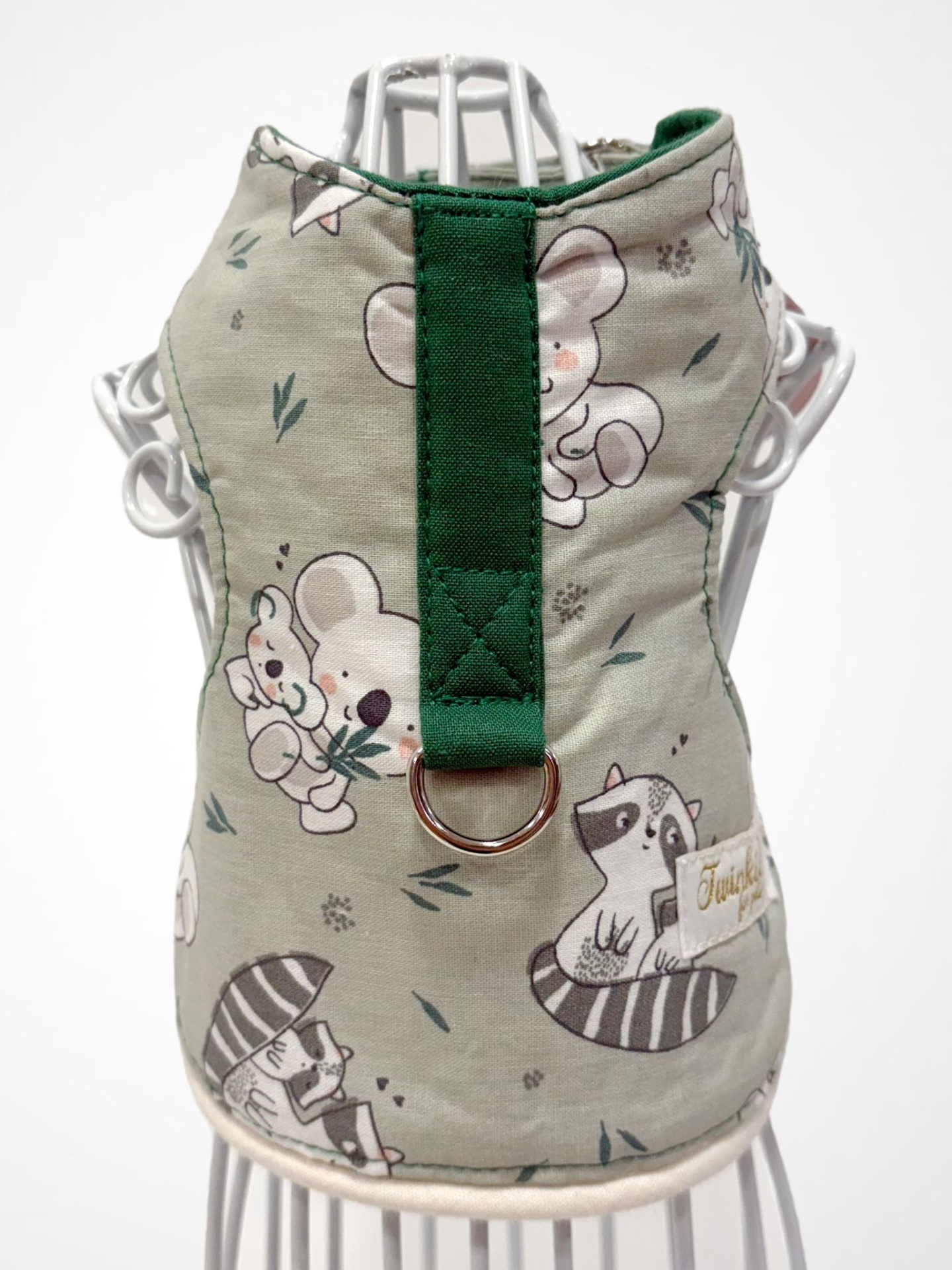 Colete para cão tecido bege com estampado de animais e fita verde com anilha metálica.