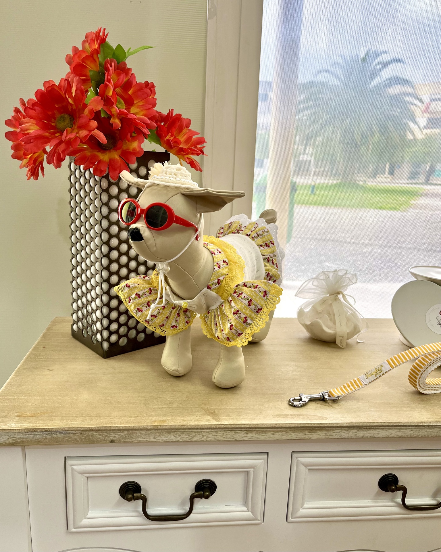 Estatueta de cão com vestido amarelo e óculos, vaso com flores vermelhas sobre móvel branco.