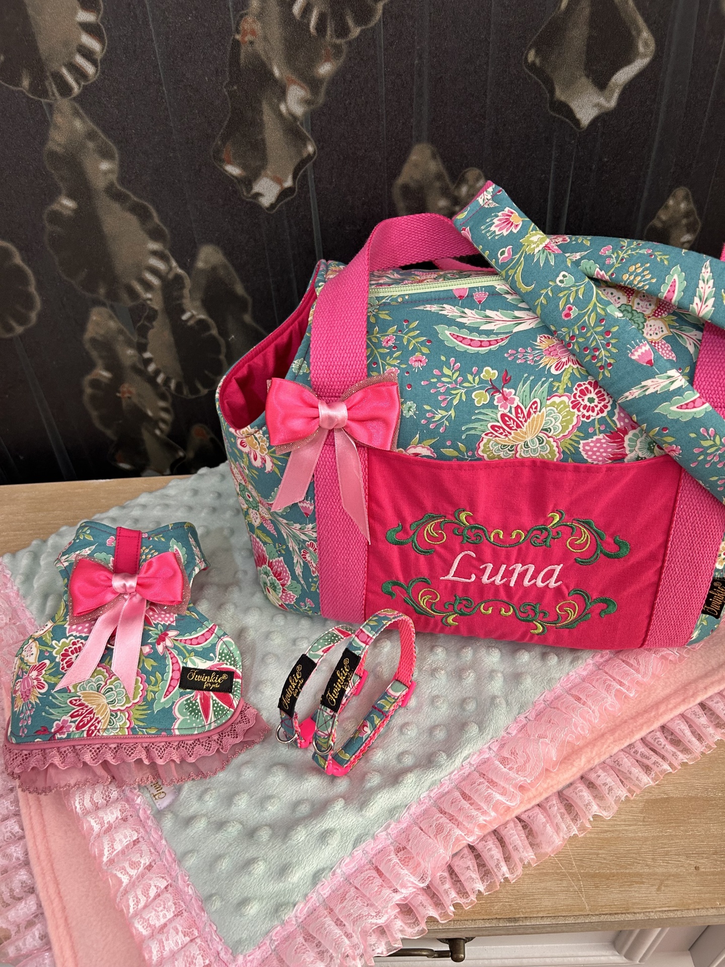Conjunto de acessórios para animal com mala floral azul e vermelho bordado, almofada rosa com fita e coleiras sobre mantas rosas