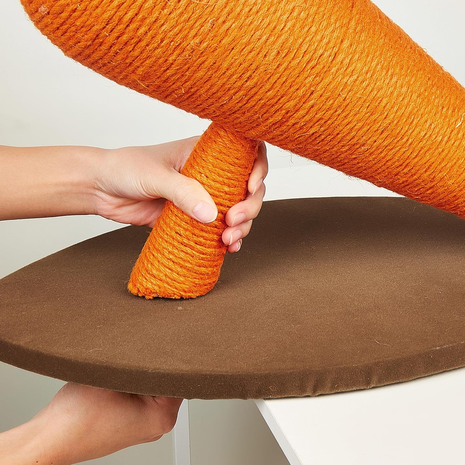 Base circular castanho com estrutura cônica de corda laranja, mãos segurando o produto