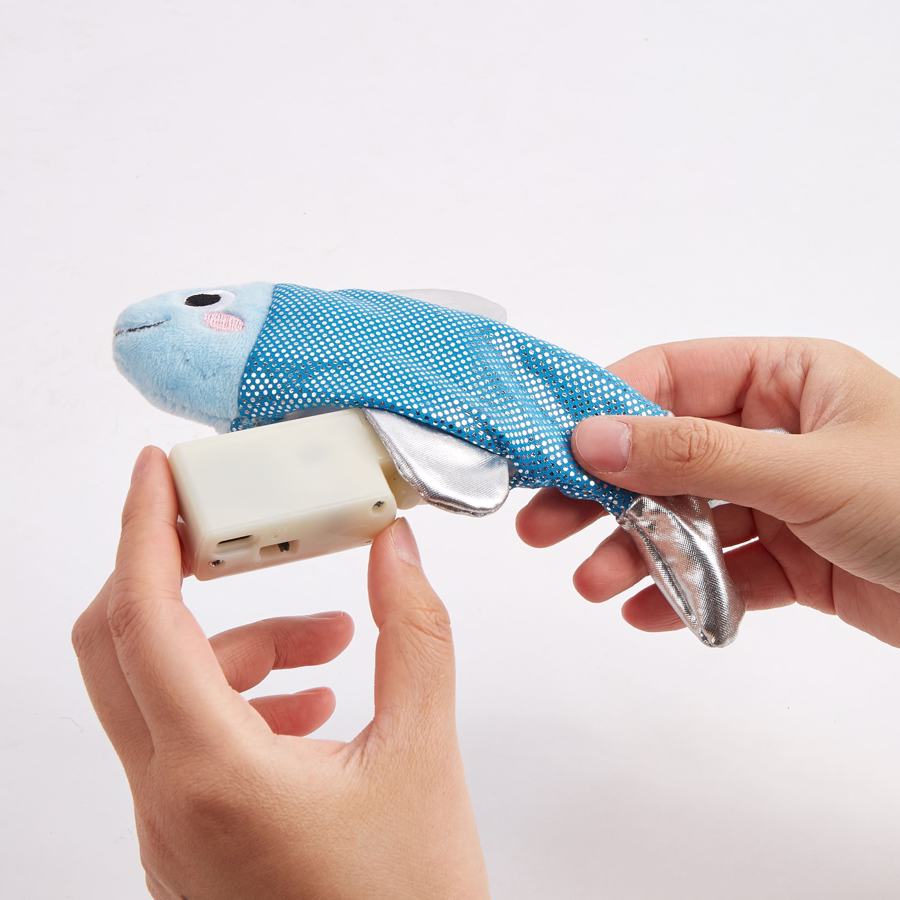 Brinquedo de pelúcia azul em forma de peixe com partes prateadas e compartimento de bateria