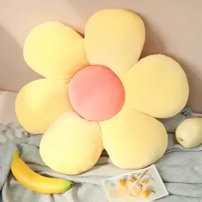 Almofada em forma de flor amarela com centro rosa, sobre tecido cinzento, com uma banana e uma maçã ao lado
