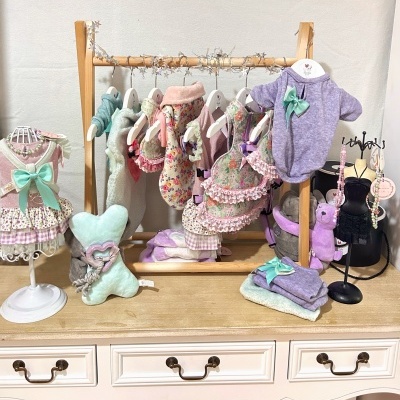 Roupinhas e acessórios florais e felpudos rosa, roxo e verde menta numa mesa branca, pendurados e expostos em manequins e cabide de madeira.