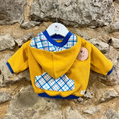 Casaco amarelo com detalhes xadrez pendurado em cabide branco contra parede de pedra