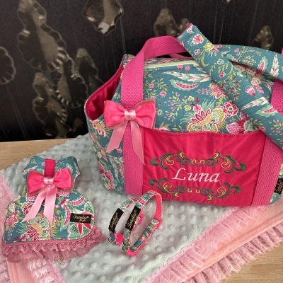 Conjunto de acessórios para animal com mala floral azul e vermelho bordado, almofada rosa com fita e coleiras sobre mantas rosas