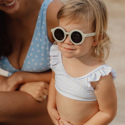 Criança com óculos de sol e top de biquíni branco com flores pequenas, junto a mulher com vestido azul de bolinhas.