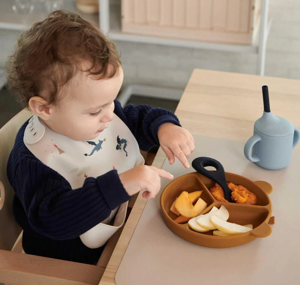Criança usando babete com desenhos a olhar para prato dividido com fruta e biberão azul
