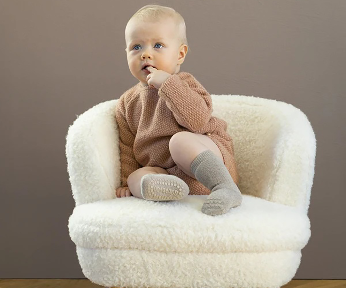 Bebé sentado numa poltrona branca felpuda com camisola castanha e meias cinza