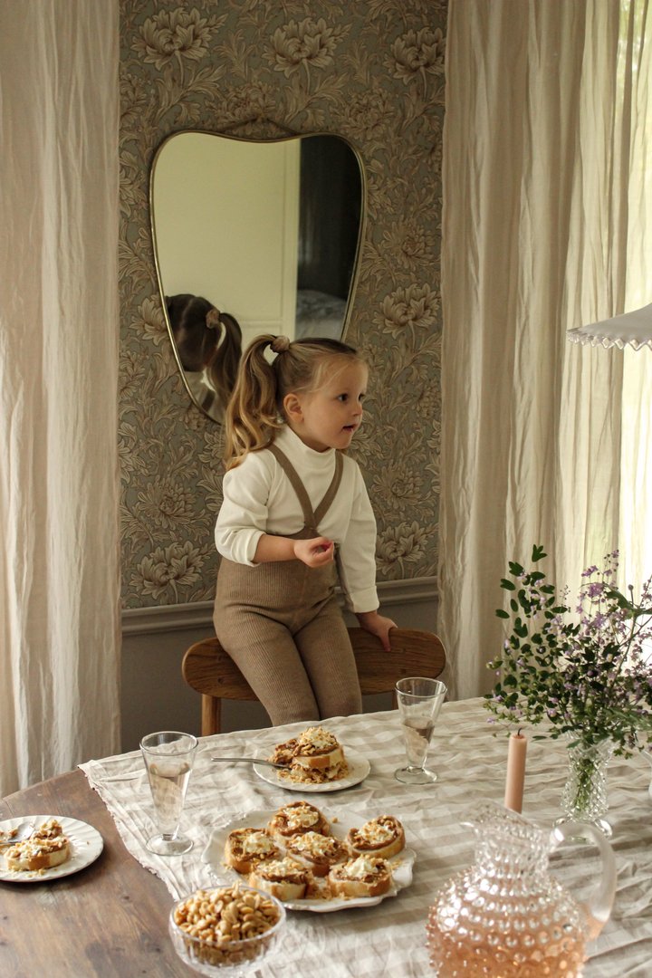 Espelho com moldura preta em parede floral atrás de criança sentada em cadeira de madeira junto a mesa com toalha bege, bolos, copos, jarra rosa e flores.
