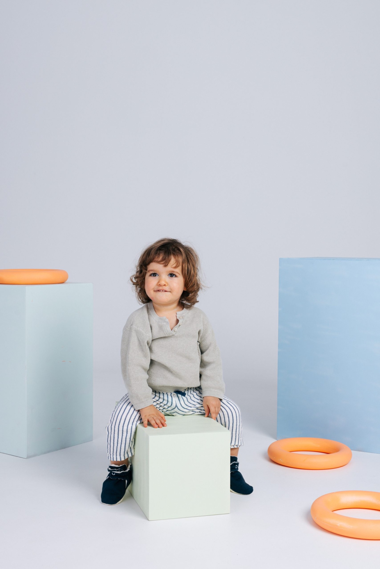 Criança sentada em caixa branca rodeada por anéis cor de laranja e caixas geométricas pastel.