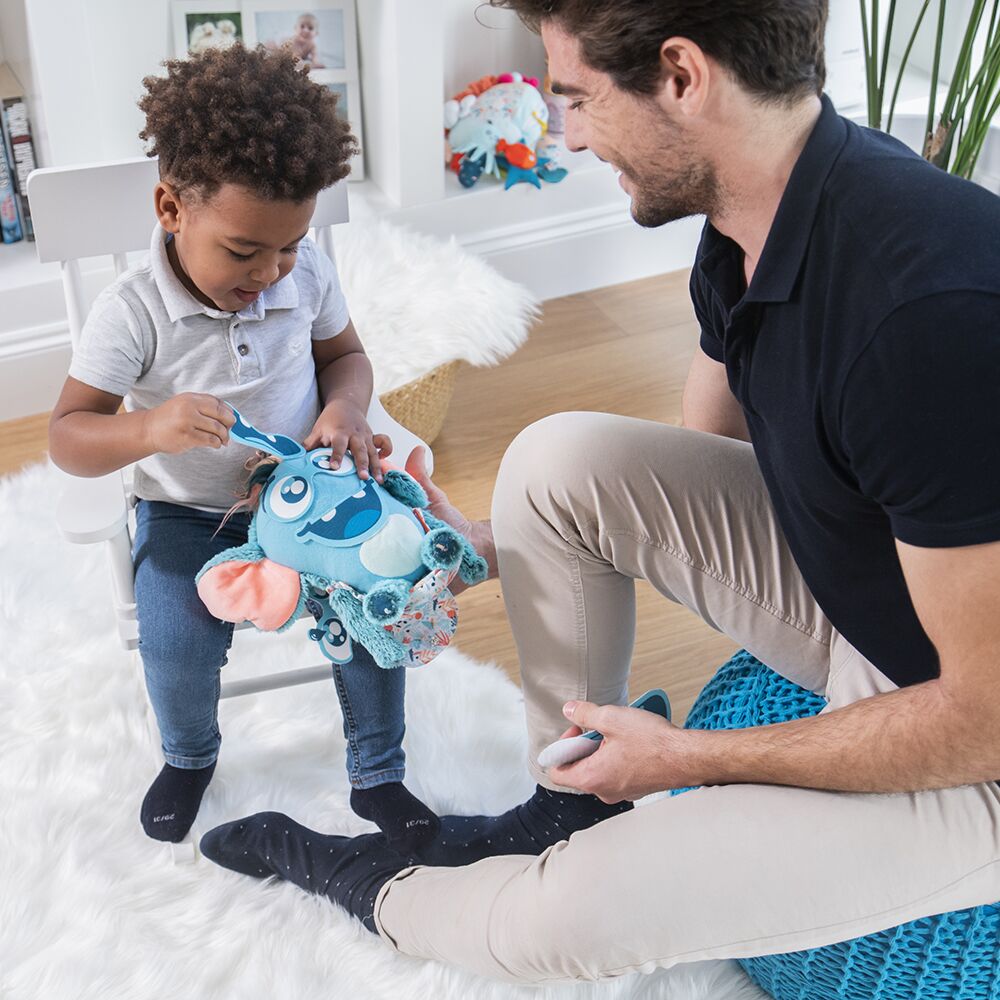 Criança com brinquedo de peluche azul e adulto sentado ao lado num ambiente interior claro