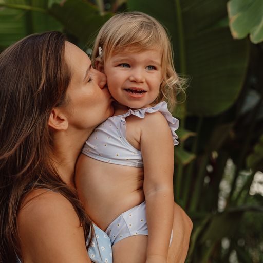 Criança pequena de biquíni azul claro abraçada por mulher adulta ao lado de planta com folhas verdes