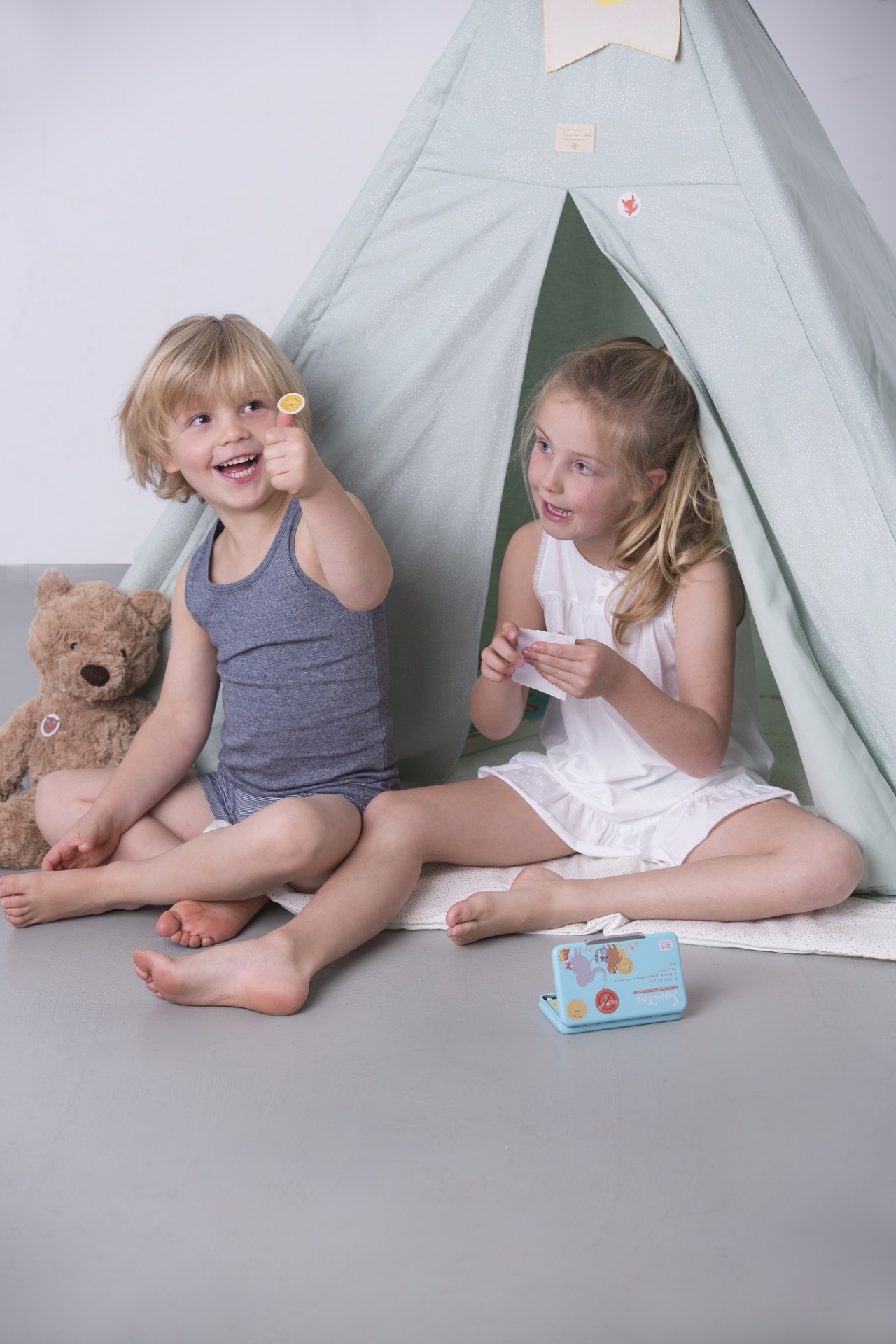 Duas crianças sentadas junto a uma tenda de tecido verde claro com um urso de peluche e um objeto azul colorido no chão