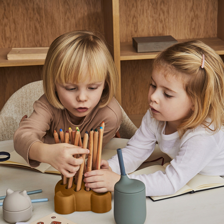 Lápis de cor de madeira organizados em suporte castanho com crianças ao redor da mesa.