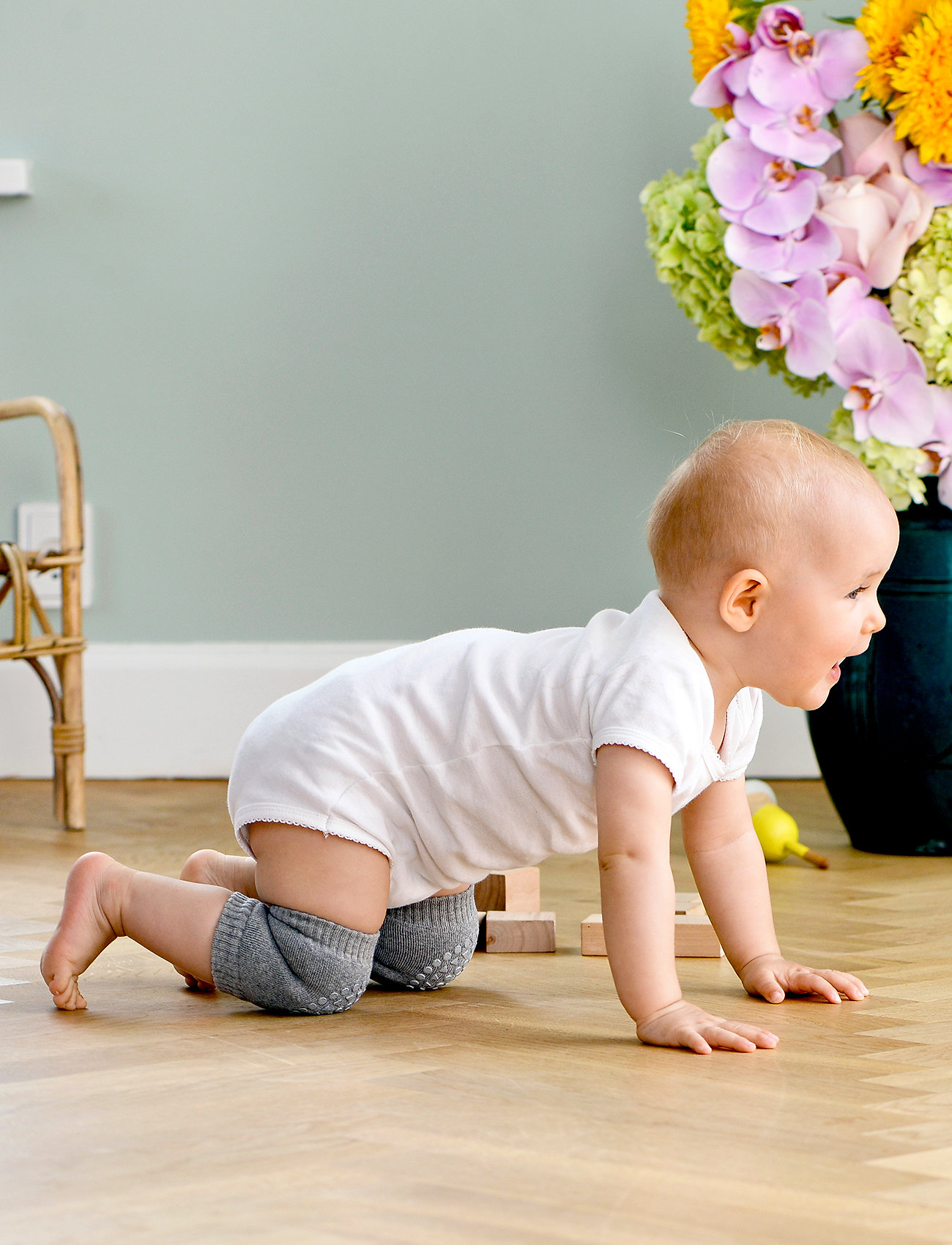 Bebé engatinhando com fato de banho branco e joelheiras cinzentas ao lado de vaso de flores e móvel de madeira.