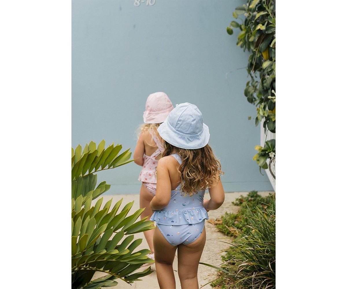Duas crianças de costas com fatos de banho e chapéus de balde, uma azul com bolinhas e outra rosa com padrão floral, ao ar livre junto a plantas.