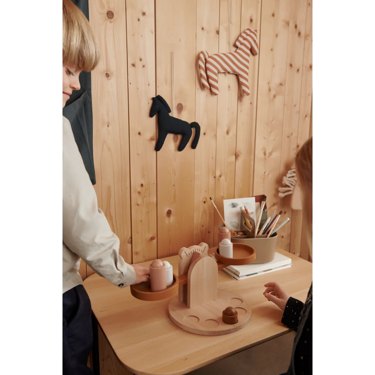 Espaço de brinquedo infantil com mesa de madeira clara, decoração de cavalos em madeira e tecido, crianças a brincar.