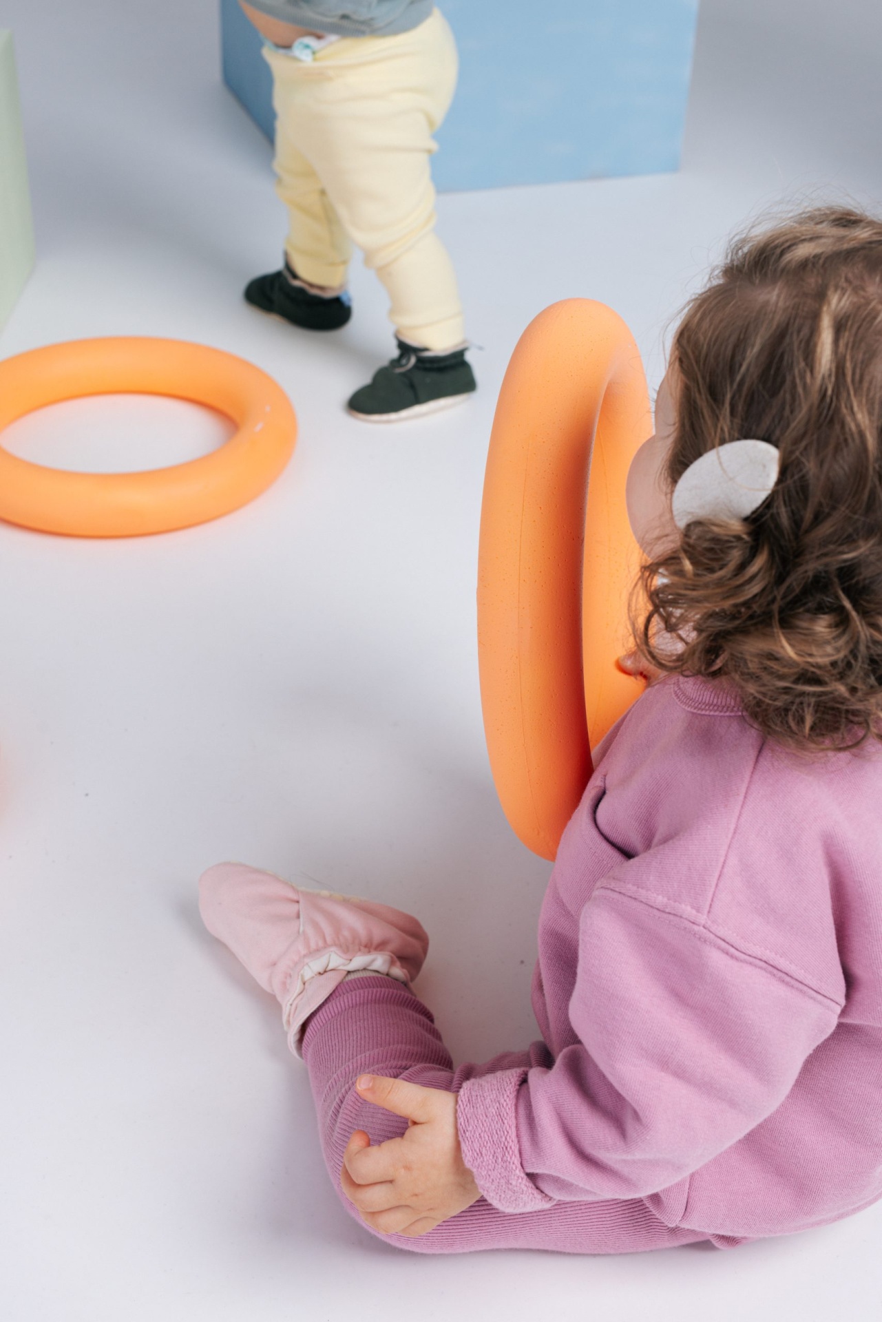 Criança com anel laranja sentado no chão e criança em pé com calças amarelas e sapatos pretos