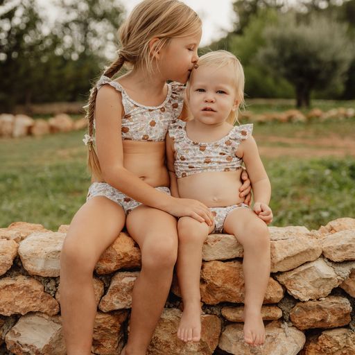 Duas crianças vestindo biquínis florais sentadas em muro de pedras ao ar livre