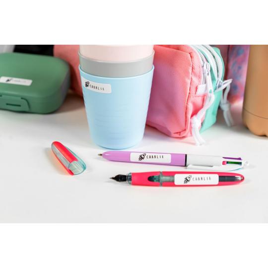 Copo azul claro, canetas coloridas com etiquetas CHARLIE, estojo rosa e estojo verde sobre mesa branca