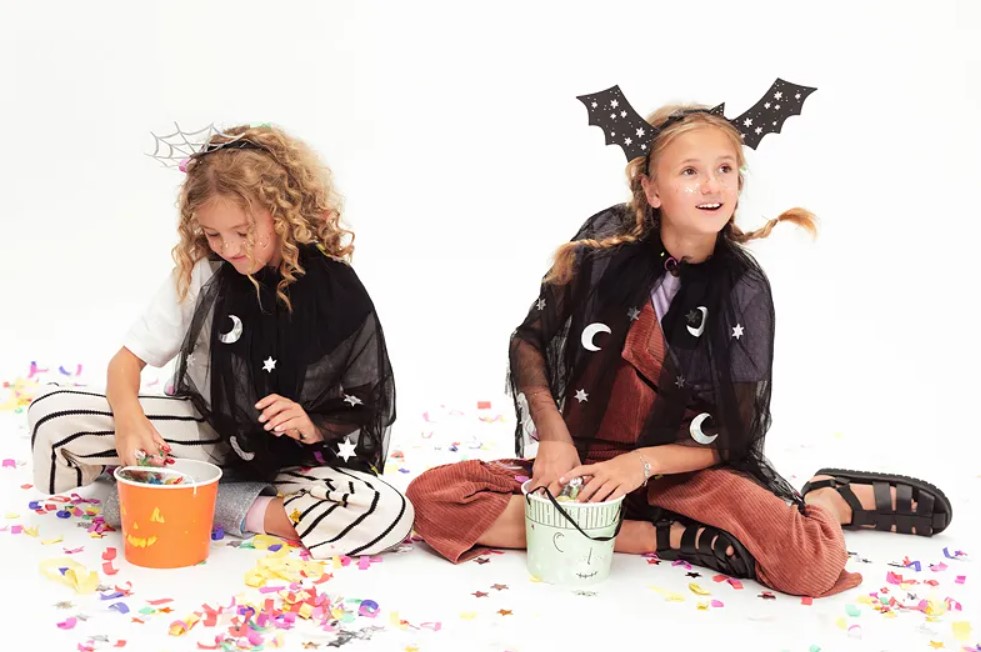 Duas crianças com mantos pretos e tiaras de Halloween, sentadas e rodeadas de confetes coloridos