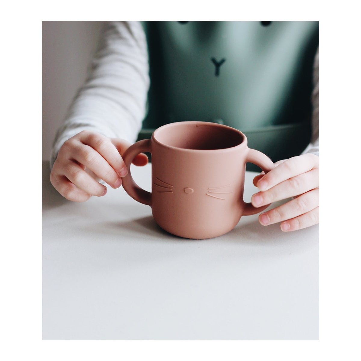 Caneca rosa clara com desenho de gato nas mãos numa mesa branca