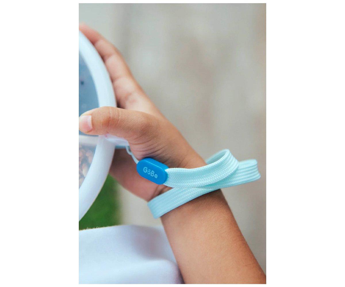 Pulseira azul claro com etiqueta azul escura G6Be no braço de criança junto a utensílio branco