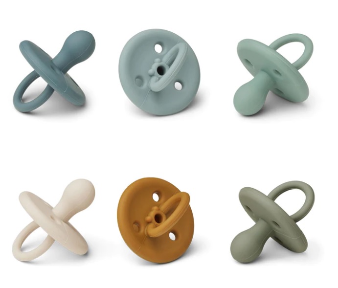 Seis chupetas de silicone em várias cores diferentes com anel, sobre fundo branco.