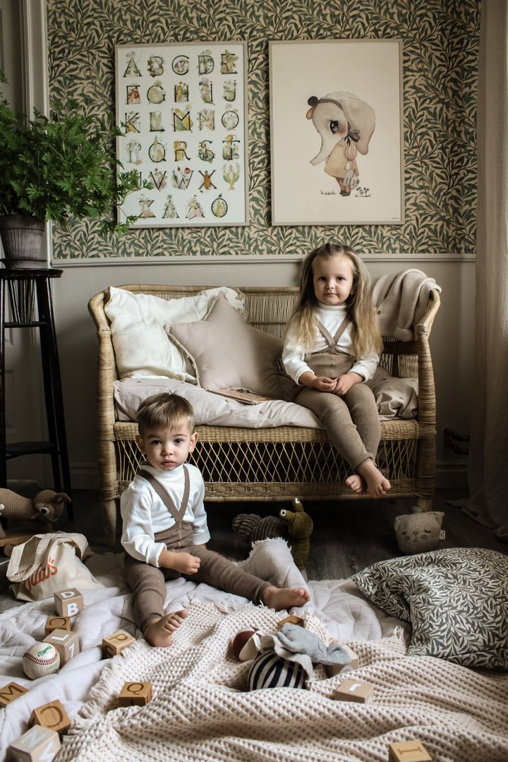 Duas crianças sentadas em sofá de vime em ambiente decorado com papel de parede floral e brinquedos no chão.