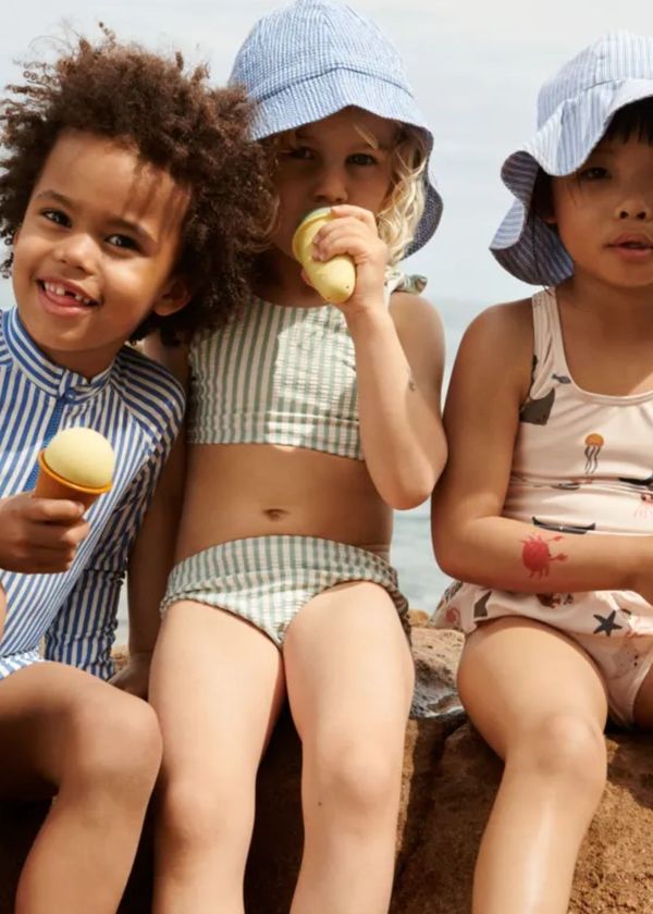 Três crianças com fatos de banho e chapéus sentadas segurando brinquedos que parecem gelados.