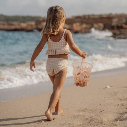 Menina com fato de banho bege e branco na praia segurando balde transparente