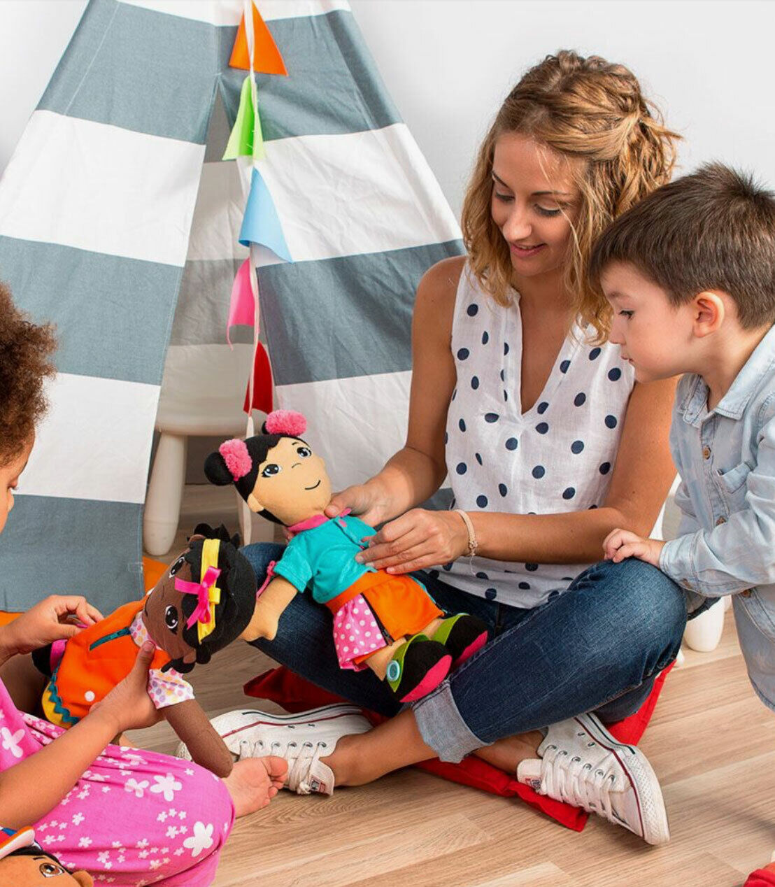 Mulher e duas crianças com bonecos têxteis coloridos junto a uma cabana listrada de tecido