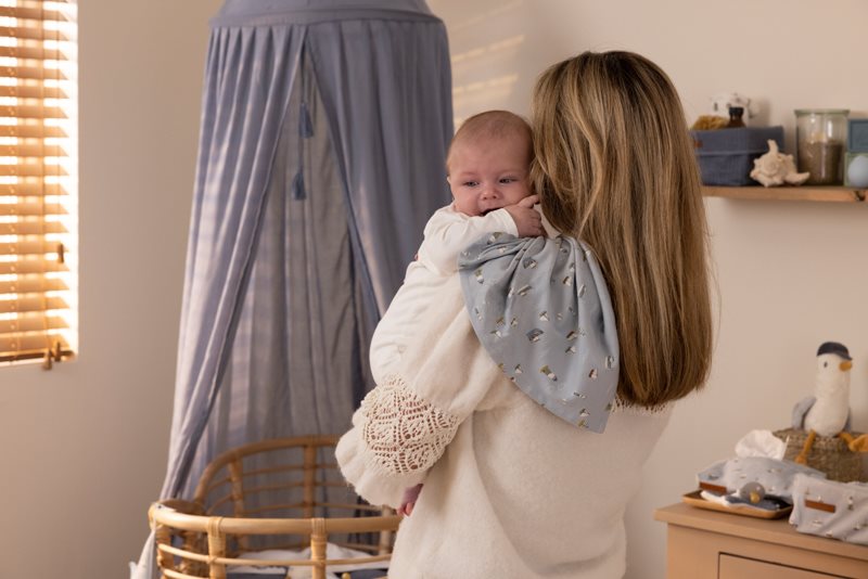 Bebé com babete cinza a ser segurado por mulher em quarto com berço redondo de madeira