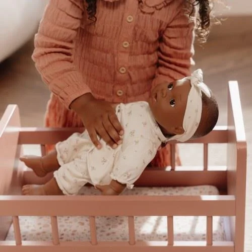 Boneca bebé com macacão branco e faixa na cabeça em berço rosa com criança