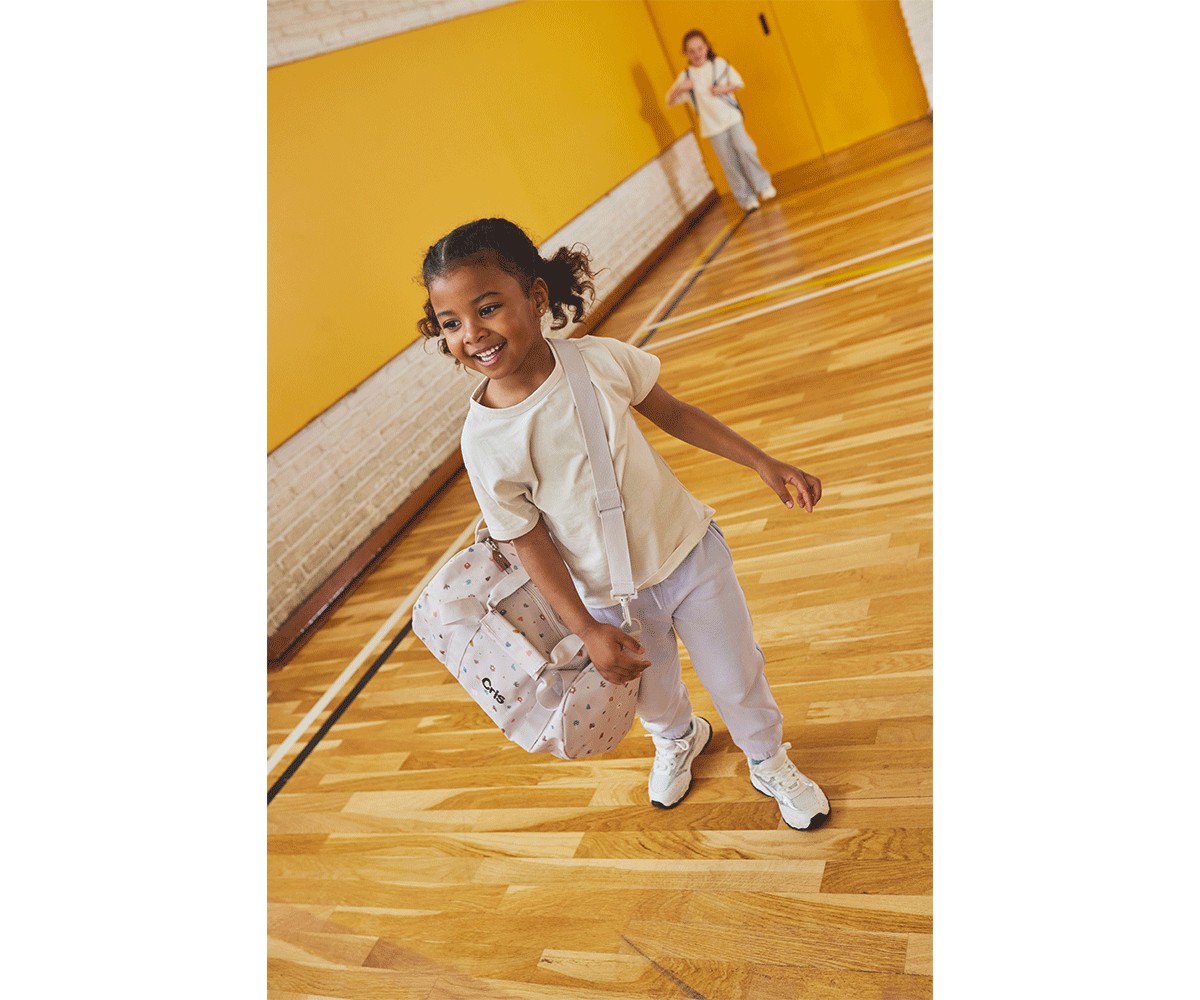 Criança sorridente com mochila branca num ginásio com parede amarela