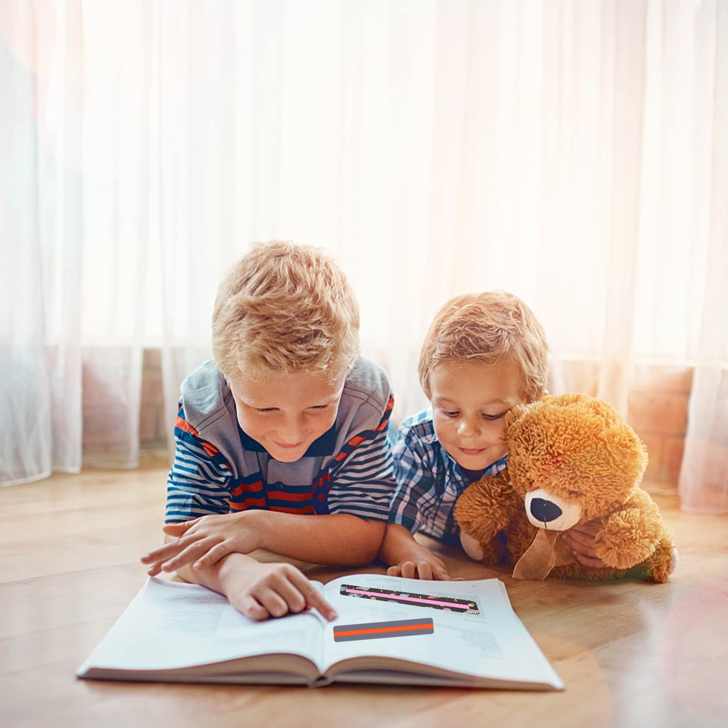 Dois rapazes loiros de camisolas às riscas deitados no chão de madeira a ler um livro, um segura um ursinho de peluche.