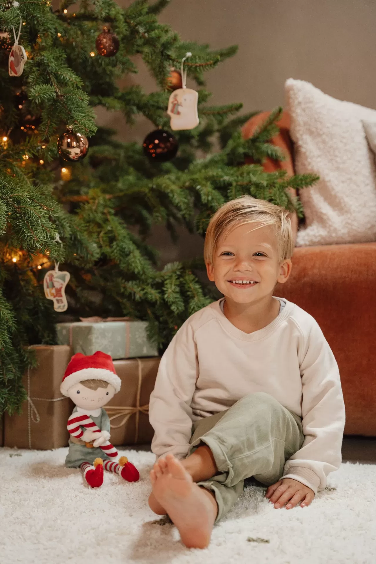 Criança sentada num tapete branco perto de árvore de Natal com enfeites e presentes