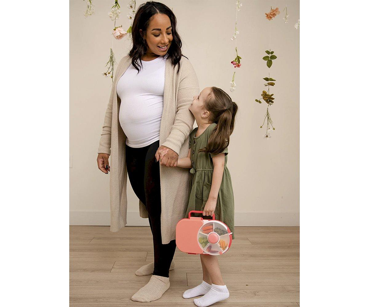 Mulher grávida com cardigan bege e menina com vestido verde num cenário com flores penduradas