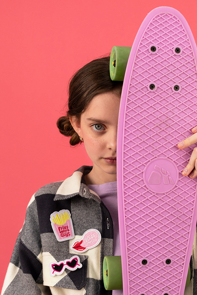 Pessoa a segurar skate rosa com rodas verdes, vestindo casaco xadrez preto e branco com emblemas coloridos