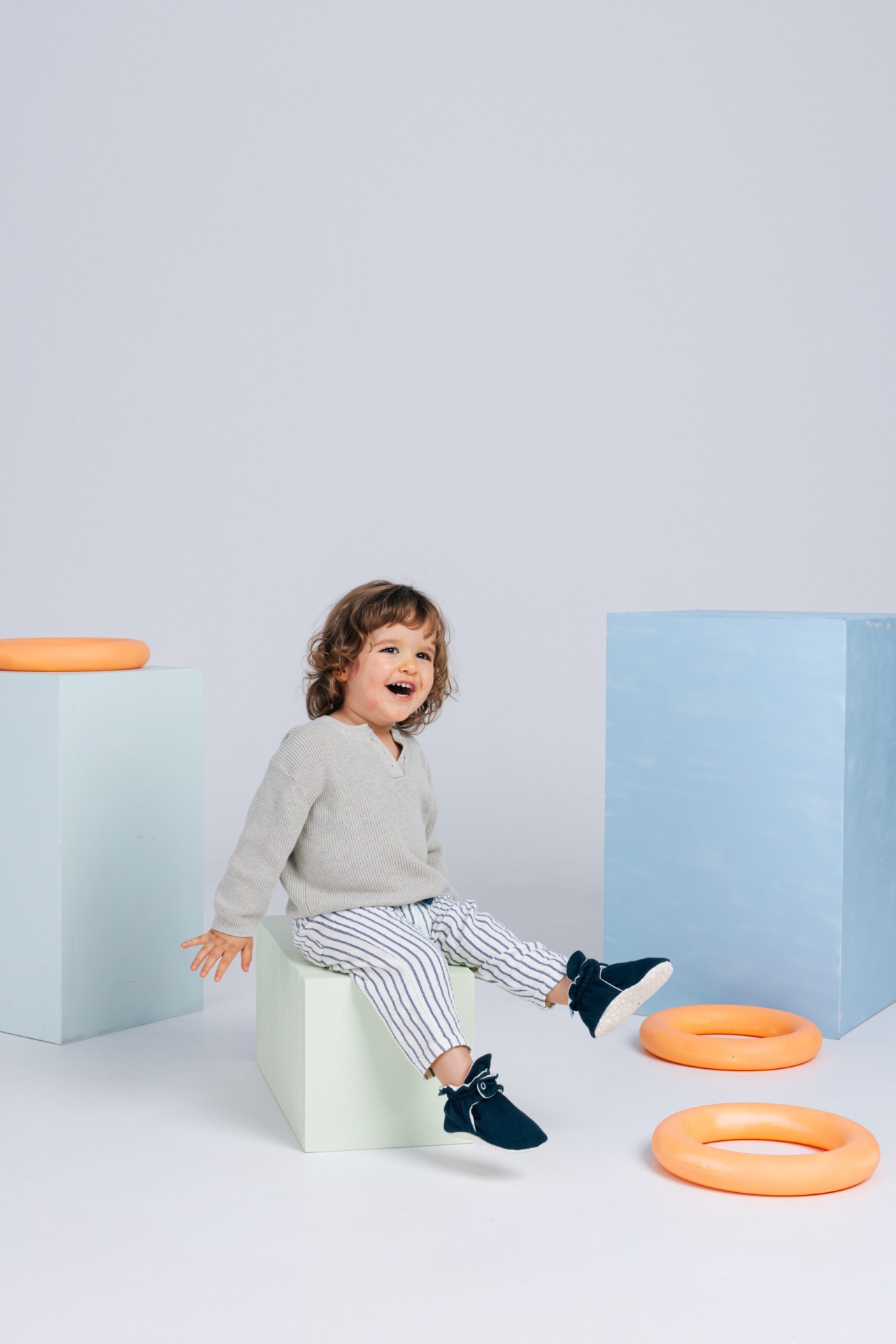 Criança feliz sentada em cubo branco com roupas claras e sapatos azuis com objetos infláveis laranjas e cubos coloridos