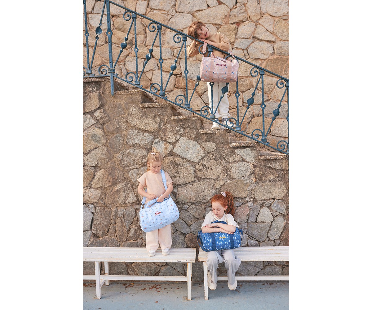 Três crianças com malas coloridas junto a uma parede de pedra e corrimão azul