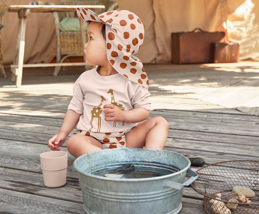 Bebé veste roupa bege com padrão de girafas e pontos castanhos, sentado na madeira com balde e copo ao lado.