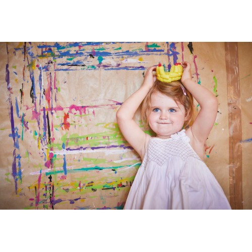 Criança com vestido branco e brinquedo amarelo na cabeça junto a parede com tinta colorida